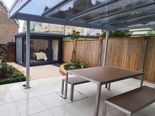 This is a beautifully designed outdoor space featuring a modern patio with sleek, polished tiles and a covered seating area. The transparent roof allows for natural light while providing protection from rain, making it a functional space for all weather conditions. The wooden fence adds privacy and warmth to the area, and the landscaped corner with greenery brings a natural touch to the setting.

The additional garden room in the background offers an ideal space for a home office, studio, or relaxation area, seamlessly blending functionality with aesthetics. If you'd like suggestions on enhancing or optimizing this area, let me know!