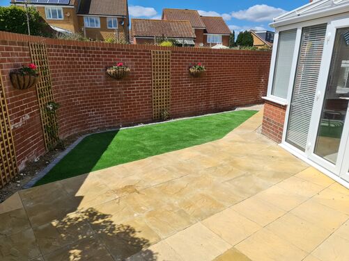 This image displays a clean and well-maintained outdoor space featuring a combination of functional and aesthetic elements. The artificial grass lawn is neatly framed with grey brick edging, providing a vibrant green accent that contrasts beautifully with the warm tones of the brick walls and the golden paving slabs.

The surrounding gravel areas add texture and drainage functionality, while the brick wall adorned with hanging planters filled with colorful flowers adds a vertical dimension and charm to the space. The conservatory on the left, with its large windows, enhances the indoor-outdoor connection, making this area an inviting extension of the home, perfect for relaxation or light gardening. The overall design exudes simplicity, low maintenance, and an eye for detail.