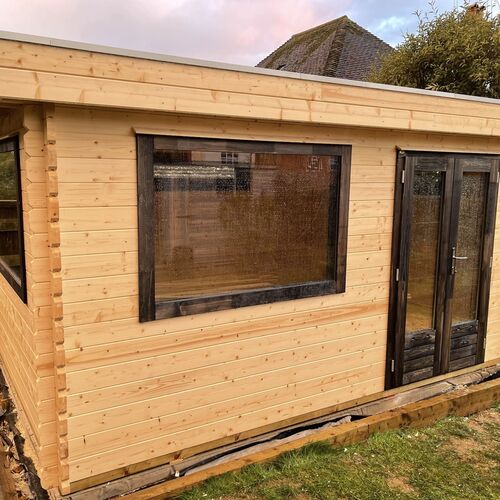 A spacious and natural wooden garden building designed and installed by Forever Timber, supplied by Lugarde. It features a light wood finish with contrasting dark-framed double glass-panel doors and large windows, providing plenty of natural light. The flat roof and rectangular design offer a versatile and functional space, perfect for use as a garden office, studio, or relaxation area.
