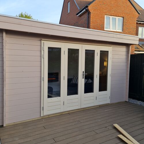 A bespoke timber garden building designed by Lugarde and created by Forevertimber. The structure features a modern flat roof and a soft, neutral painted exterior. The front is adorned with elegant full-length glass double doors and windows, offering ample natural light. It is situated on a composite deck with clean, modern surroundings, making it a perfect space for relaxation or as a stylish garden retreat.