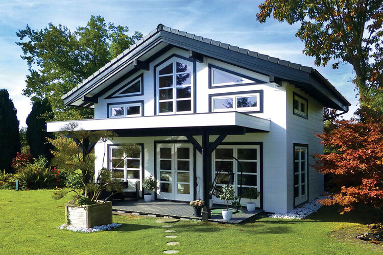 This modern accommodation is a two-story house with a black roof, white walls, and large windows. The front features a small porch adorned with potted plants. It is nestled in lush green lawns, among trees and colorfull foliage, under a clear blue sky.