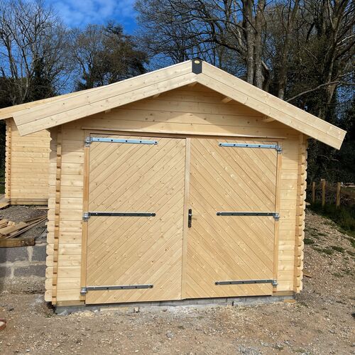 "High-quality timber garage with a pitched roof and natural wood finish, constructed with durable interlocking logs. Features large double doors with a diagonal chevron design, reinforced by robust metal hinges and a central locking mechanism for security. The structure sits on a sturdy concrete base, ensuring long-lasting stability. Surrounded by a natural landscape, this garage offers a functional and aesthetically pleasing solution. Supplied by Lugarde and expertly designed and installed by Forever Timber, it is a perfect blend of craftsmanship and practicality."