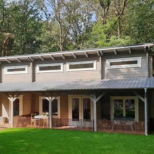Contemporary two-story timber house with a flat-sloped roof and a stylish gray finish. The design features large rectangular windows on both floors, with narrow clerestory windows on the upper level providing ample natural light. The lower level includes a covered porch supported by clean-lined timber posts, creating a cozy outdoor seating area. Surrounded by lush greenery and a well-manicured lawn, the house seamlessly blends with its natural environment. Supplied by Bertsch Holzbau and expertly designed and installed by Forever Timber, this home combines modern architectural elegance with high-quality craftsmanship.