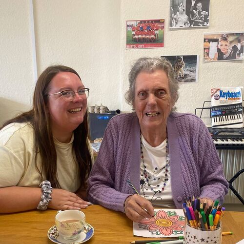 Elderly lady smiling next to her carer, whilst sat at the table colouring in at Your Care's Dementia Day Club in Hanham, Bristol