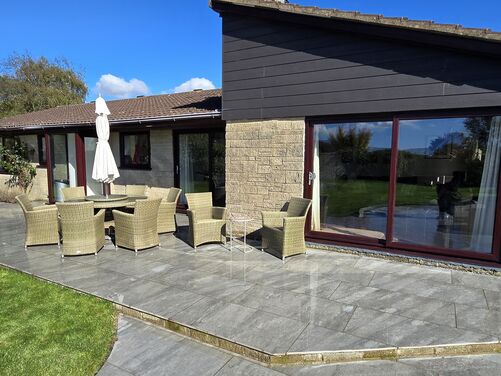 The image features a spacious and elegant outdoor patio area adjoining a modern home with large glass windows and sliding doors. The patio is paved with shiny gray tiles, giving it a polished and contemporary look. A set of wicker chairs and a round table with an umbrella provide a comfortable seating area for outdoor dining or relaxation. The patio extends into a lush, well-maintained lawn, creating a seamless connection between the hardscaped and green spaces. The house's stone and wood exterior enhances the rustic yet modern aesthetic, while the clear blue sky and natural surroundings create a welcoming and serene atmosphere.