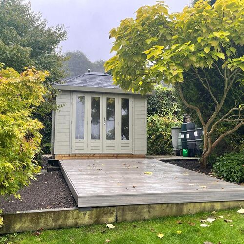 A bespoke timber garden building supplied by Lugarde and designed and installed by Forevertimber. This cozy structure features a light sage-green painted exterior with glass-panelled double doors and a matching window design, allowing natural light to fill the interior. The building is topped with a slate-effect pitched roof, adding a classic touch. Positioned on an elevated wooden deck surrounded by lush greenery, it creates a serene and inviting atmosphere. This garden retreat is ideal for relaxation, as a home office, or a hobby space, blending seamlessly with its natural surroundings.