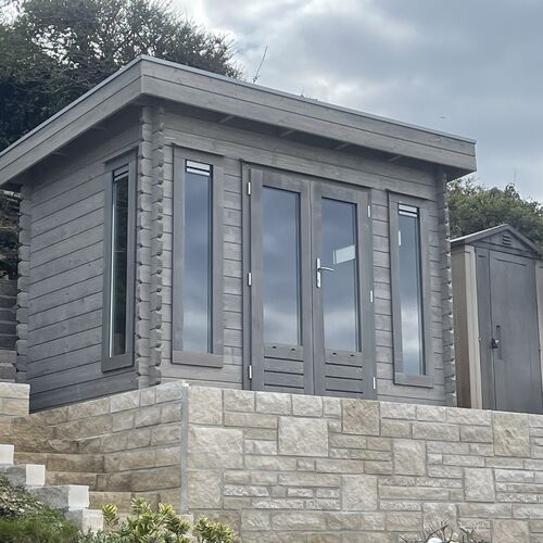 A bespoke timber garden building supplied by Lugarde and designed and installed by Forevertimber. This compact and stylish structure features a flat roof and a modern grey-stained timber exterior. The design includes full-length glass-panelled double doors and tall, narrow side windows, offering ample natural light and a contemporary aesthetic. The building is elevated on a beautifully constructed stone platform with steps, enhancing its prominence and accessibility. It is perfect for use as a garden office, studio, or retreat, seamlessly blending functionality with a modern design.
