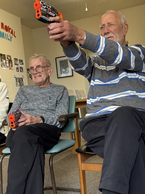 Two seated guests hold foam dart blasters, with one taking aim during a game at Your Care Dementia Day Club in Hanham.