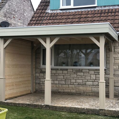 A bespoke garden gazebo supplied by Lugarde and designed and installed by Forevertimber. This structure features a light-painted timber frame with sturdy posts and decorative cross-bracing for support. The flat roof provides a modern aesthetic, while the back wall is constructed with natural wood paneling, offering privacy and additional protection from the elements. The gazebo is positioned adjacent to a house with stone and brickwork, seamlessly integrating into the existing architecture. The covered area provides a functional and sheltered space, ideal for outdoor seating or storage, blending practicality with stylish design.