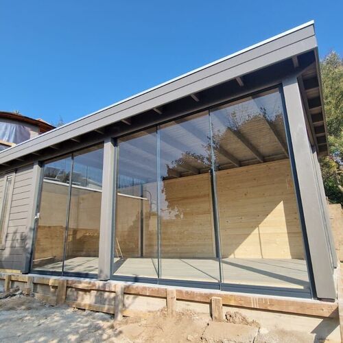 A bespoke timber garden building designed by Lugarde and created by Forevertimber. This modern structure features a sleek, asymmetrical slanted roof and expansive glass walls that provide panoramic views and abundant natural light. The interior showcases natural timber paneling, giving it a warm and inviting feel. The open, minimalist design makes it ideal for a garden studio, office, or relaxation space. It is elevated on a sturdy base, seamlessly blending contemporary design with practicality.