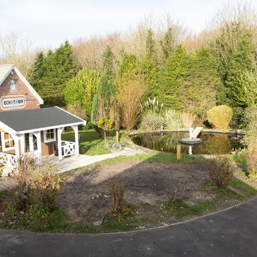 A charming and rustic garden building designed and installed by Forever Timber, supplied by Lugarde. The structure features a classic pitched roof with a covered porch, adorned with white railings, giving it a cozy, cabin-like appearance. It sits beside a beautifully landscaped pond with a fountain, surrounded by lush greenery, creating a serene and picturesque outdoor setting. Perfect for a relaxing retreat or unique garden addition.