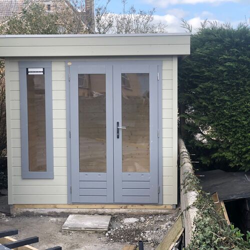 A compact and modern garden building designed and installed by Forever Timber, supplied by Lugarde. It features a light cream exterior with grey accents, including double glass-panel doors and a tall vertical window, providing a bright and inviting space. Its flat roof and sleek design make it an ideal addition to smaller gardens, perfect for use as an office, studio, or relaxation area.