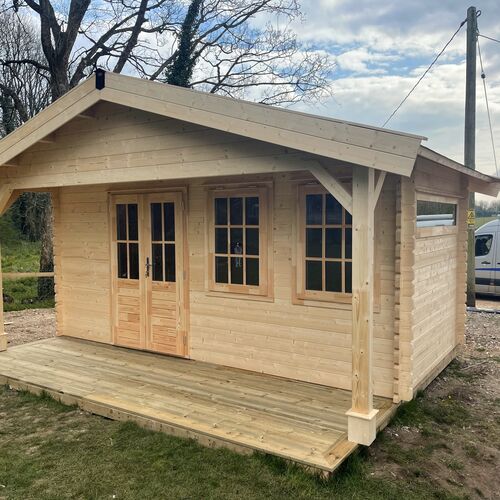 "Beautiful timber cabin with a pitched roof and extended front porch, crafted from natural wood with a light finish. Features include double French doors with glass panels and matching windows, allowing plenty of natural light inside. The porch is supported by elegant timber posts with decorative braces, creating a welcoming and functional outdoor space. Set on a raised wooden platform, this cabin exudes rustic charm and versatility. Supplied by Lugarde and skillfully designed and installed by Forever Timber, it’s a perfect solution for a garden retreat or multi-purpose outdoor building."