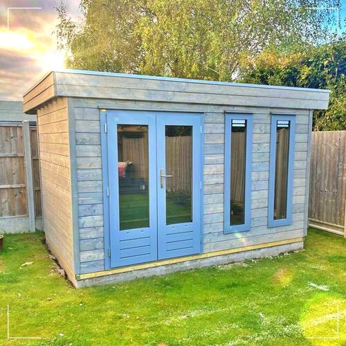 "Modern timber garden building with a flat roof, supplied by Lugarde and expertly designed and installed by Forever Timber. The structure features a sleek grey exterior with clean horizontal cladding, complemented by a set of double glass doors and tall vertical windows for maximum natural light. The building is raised slightly on a sturdy base, set on a well-maintained lawn and surrounded by wooden fencing for privacy. Ideal for use as a garden office, studio, or relaxation space, this structure combines contemporary aesthetics with high-quality craftsmanship to enhance any outdoor area."