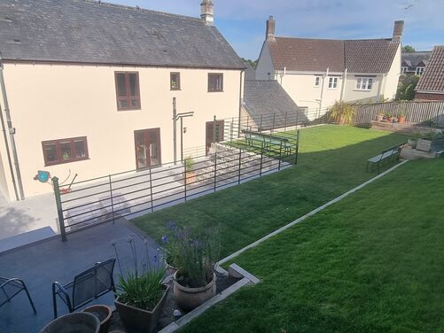 The image shows a spacious and well-organized outdoor area of a property. A modern home with a cream facade and slate roof serves as the focal point. The area features a neatly maintained lawn on a gentle slope, bordered by retaining walls and fencing, creating defined levels.

The lower section includes a paved patio with light gray tiles, surrounded by black railings for safety. This space connects seamlessly to the house through glass doors, making it ideal for outdoor dining or entertaining. The upper lawn features raised planters with flowers and shrubs, adding greenery and natural beauty. In the background, neighboring homes and gardens are visible, blending into the peaceful and landscaped surroundings. The layout maximizes usability while maintaining an open and inviting aesthetic.