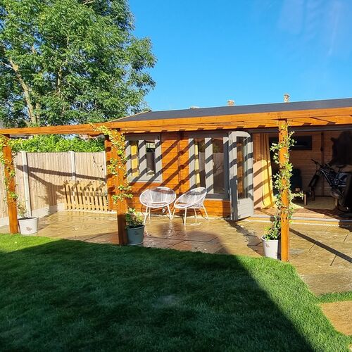 Stylish timber garden building with a flat roof, complemented by an adjoining pergola covered in climbing plants for a natural and inviting outdoor space. The structure features large glass doors and windows, allowing plenty of natural light into the interior. The surrounding area includes a beautifully paved patio and a lush green lawn, creating a perfect setting for relaxation or entertaining. Furnished with modern outdoor seating and potted plants, this garden retreat, designed and installed by Forever Timber, combines practicality and elegance, making it an ideal addition to any outdoor living area.