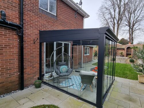 The image features a modern outdoor conservatory attached to a brick house. The conservatory is constructed with black metal framing and glass walls, providing a clear view of the garden while offering a sheltered seating area. Inside, there is a cozy setup with a hanging egg chair adorned with cushions, a small table, and additional seating with a patterned outdoor rug adding a touch of color and texture.

The surrounding area includes light-colored paved stone flooring that transitions into a green lawn. In the
