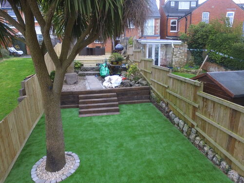 The image shows a backyard or garden area with a mix of artificial turf and landscaped features. In the foreground, there's a large tree surrounded by decorative stones and bricks, and the ground is covered with bright green artificial grass. A set of wooden steps leads up to a higher level with a patio area and some plants, partially covered with gravel and wooden decking. The space is enclosed by wooden fencing, with neighboring brick houses visible in the background.