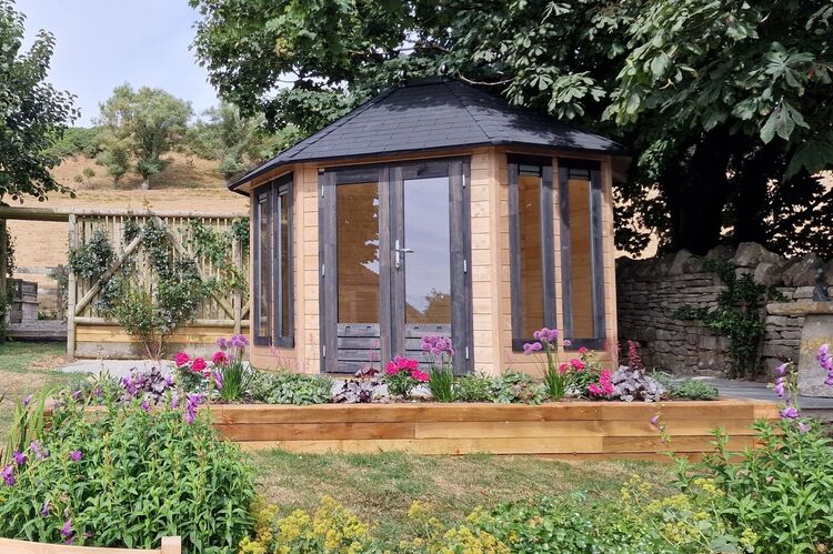 A small octagonal wooden garden room with large windows is surrounded by colorful flowers and plants. It sits on a stone base, under the shade of trees, with a hillside and wooden trellis in the backdrop. This bespoke Lugarde retreat harmoniously blends nature and elegant design.