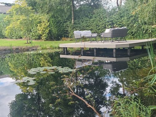 The image captures a tranquil and scenic outdoor space featuring a serene pond surrounded by lush greenery. A wooden deck extends over the edge of the pond, furnished with elegant outdoor seating, including cushioned chairs and a table, creating an inviting spot for relaxation or socializing.

The pond reflects the surrounding trees and sky, enhancing the peaceful atmosphere. Water plants, including reeds and lily pads, add natural charm and biodiversity. In the background, a well-manicured lawn leads to a modern house with large windows, blending contemporary architecture with the natural beauty of the landscape. The setting is idyllic, offering a perfect retreat for enjoying nature.