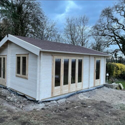 A bespoke timber garden building designed by Lugarde and crafted by Forevertimber. This structure features a pitched roof with a classic design and a light-painted exterior accented by natural wood-framed windows and doors. The front facade boasts multiple full-length glass doors, providing ample natural light and creating a bright, open atmosphere. Its spacious layout makes it versatile for various uses, such as a garden room, home office, or hobby space. The surrounding area appears to be under landscaping preparation, emphasizing its potential as a stunning garden feature.
