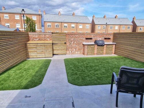 This backyard features a contemporary and minimalist design, blending clean lines with functional elements. The space includes artificial grass sections framed by paved walkways, surrounded by horizontal wooden slat fencing for privacy and style. The focal point is a brick-built outdoor kitchen or barbecue area, creating an ideal setup for entertaining and outdoor dining.

The balance of greenery and hardscaping provides a low-maintenance yet inviting outdoor space.