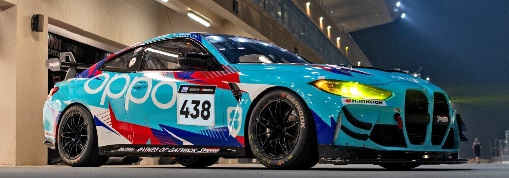 BMW M4 GT4 with blue motorsport wrap and sponsor graphics next to race track at night.