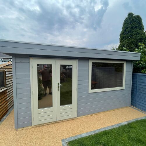 A bespoke timber garden building supplied by Lugarde and designed and installed by Forevertimber. This structure features a modern flat roof and a soft grey-painted timber exterior, complemented by white-framed double glass doors and a large rectangular window. The combination of doors and windows provides ample natural light, making it an ideal space for a garden office, studio, or relaxation area. The building is set on a light resin-bound pathway with neat landscaping, creating a clean and contemporary look that blends seamlessly with the garden surroundings.