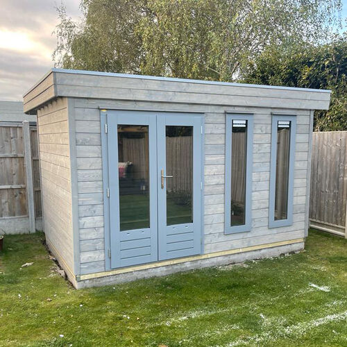 A bespoke timber garden building supplied by Lugarde and designed and installed by Forevertimber. This contemporary structure features a flat roof and a soft grey finish, giving it a sleek and modern look. The front facade includes full-length glass-panelled double doors and two tall, narrow windows, providing excellent natural light and a sense of openness. The minimalist design and clean lines make this building a stylish addition to any garden, ideal for use as a home office, studio, or relaxation space. Set on a neatly maintained lawn and enclosed by fencing, it integrates seamlessly into the outdoor environment.