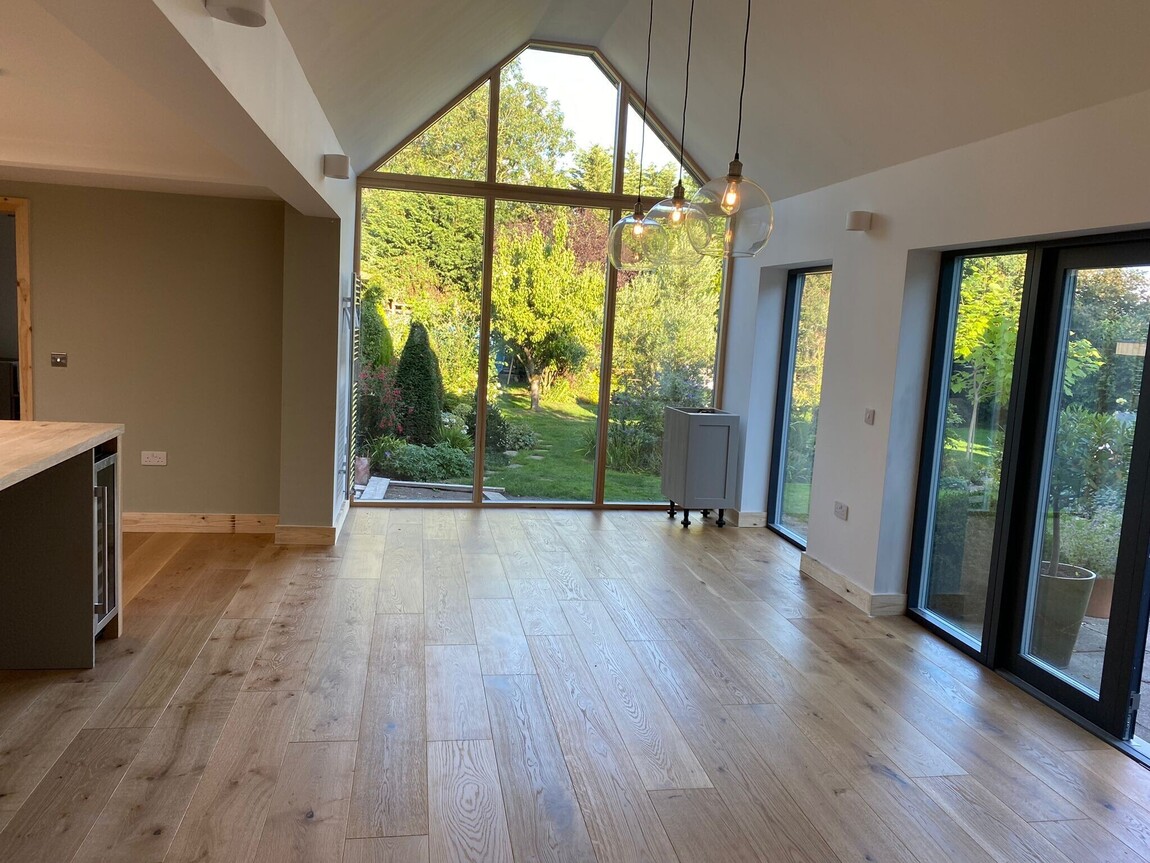Bright and airy open-plan living space featuring a vaulted ceiling and stunning floor-to-ceiling glass windows that offer seamless views of the lush garden. The natural wood flooring and modern pendant lighting create a warm and inviting atmosphere, perfect for blending indoor and outdoor living