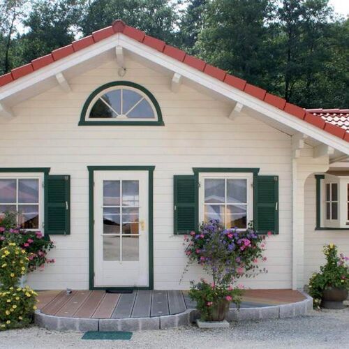 Charming single-story timber house with a red-tiled pitched roof and a bright white exterior. The house features green-framed windows with matching shutters, adding a pop of color and a traditional touch. A central arched window above the entrance door provides an elegant focal point, while the front door includes glass panels to allow natural light inside. The entrance is framed by vibrant flower beds and potted plants, creating a welcoming and picturesque setting. Supplied by Bertsch Holzbau and beautifully designed and installed by Forever Timber, this house combines timeless appeal with expert craftsmanship.