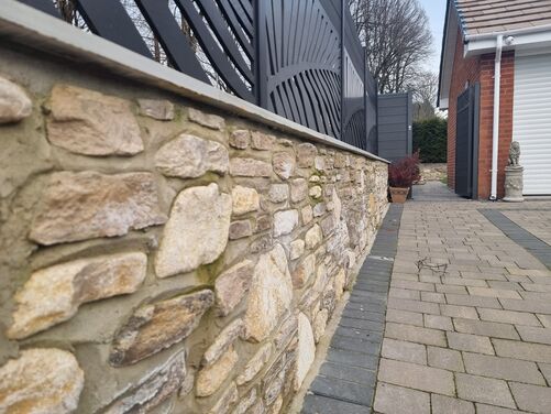 The image showcases a close-up view of a beautifully crafted garden wall made of natural stone, offering a rustic yet elegant appearance. The wall is topped with a smooth, light-colored coping stone for a finished look. Above the wall, decorative black metal panels add a modern touch, featuring an intricate sunburst pattern that provides both style and privacy.

The adjacent pathway is made of neatly arranged paving stones bordered by darker bricks, enhancing the clean and structured design. In the background, a red brick house and garage are partially visible, along with potted plants and a decorative statue, creating a cohesive and well-maintained outdoor space.