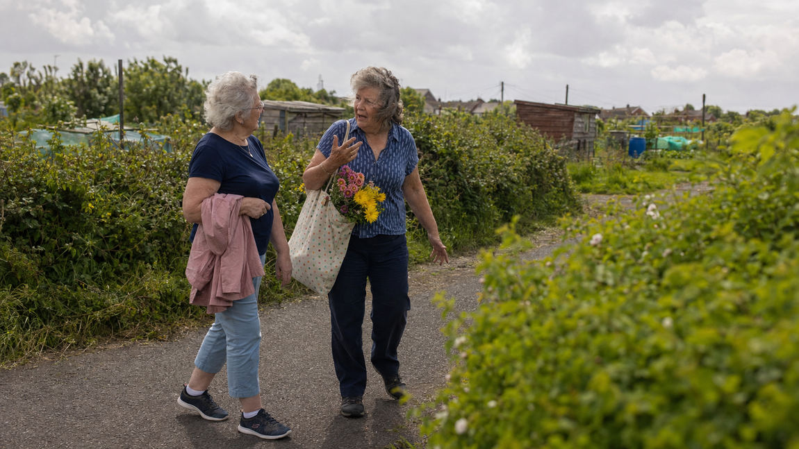 Two older female friends walking and talking together beside their local allotments on a summer afternoon.