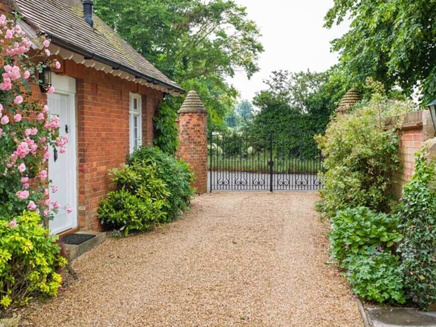 Charming cottage entrance with a gravel driveway framed by lush greenery and blooming flowers. The red brick walls and decorative wrought iron gate add a timeless, rustic touch, leading to a serene countryside setting