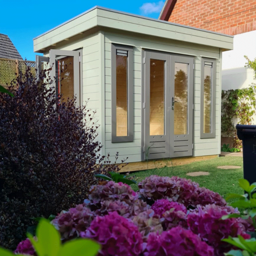 A bespoke timber garden building supplied by Lugarde and designed and installed by Forevertimber. This elegant structure features a flat roof and a light sage green exterior with contrasting dark-framed full-length glass doors and windows. The design emphasizes natural light and a modern aesthetic, making it an excellent choice for a garden office, studio, or retreat. The surrounding landscaping includes vibrant pink hydrangeas and well-maintained greenery, enhancing the building's charm and integration into the garden. Its compact yet stylish design complements both residential and natural environments.