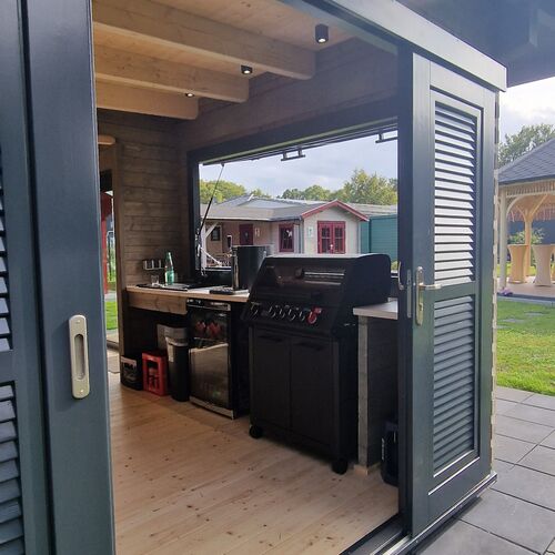 Interior view of a modern timber pavilion designed for outdoor cooking and entertainment, featuring a built-in kitchenette and a high-quality barbecue grill. The space is enclosed with sliding glass doors and louvered panels, offering both ventilation and weather protection. The natural wood interior with exposed beams creates a warm and inviting atmosphere. Practical storage cabinets and counter top space provide functionality, while the open design allows for seamless interaction with the outdoor area. Designed and installed by Forever Timber, this pavilion blends contemporary style with expert craftsmanship, making it a perfect setting for outdoor gatherings.