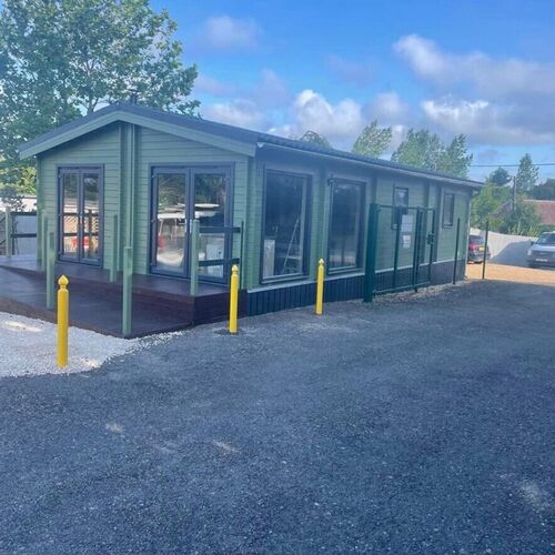 Modern timber house with a pitched roof, featuring a sleek green exterior complemented by large glass windows and sliding doors that allow abundant natural light. The structure is elevated on a dark wood deck, creating a clean and practical base with ample space for outdoor activities. Surrounded by a gravel driveway and secured with yellow bollards for safety, this house is designed for both functionality and style. Supplied by Lugarde and expertly designed and installed by Forever Timber, it offers a contemporary living solution with durable materials and meticulous craftsmanship.