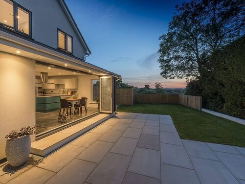 This photo showcases a stunning integration of indoor and outdoor living. The fully open bi-fold doors create a seamless connection between the kitchen and dining area and the outdoor patio, perfect for entertaining or enjoying warm evenings. 

The lighting highlights the modern design, with recessed ceiling lights enhancing the indoor area and subtle outdoor lighting ensuring the patio is inviting after sunset. The use of large, neutral-colored patio slabs complements the grass lawn, creating a balanced and harmonious outdoor space. 

The view beyond the garden adds an open, expansive feeling, enhancing the property's ambiance. The setup is both practical and aesthetically pleasing, providing versatility for various uses.