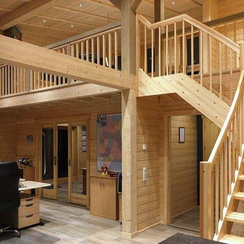 Spacious and beautifully crafted timber interior featuring a two-story layout with natural wood paneling throughout. The design includes an open mezzanine level with elegant wooden railings, accessible via a wide staircase, providing a blend of functionality and aesthetic appeal. The ground floor features a work area with desks and cabinetry, offering ample storage and organization options. Large glass-paneled doors allow natural light to fill the space, complementing the warm tones of the wood. Designed and installed by Forever Timber, this interior highlights exceptional craftsmanship, making it perfect for residential or commercial use.