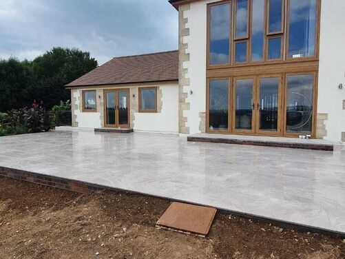 A modern house with a large, newly constructed patio featuring polished light grey tiles. The house has large glass windows and wooden-framed doors, with stone accents on the exterior. The surrounding area includes a landscaped garden with plants and an unfinished dirt section in the foreground