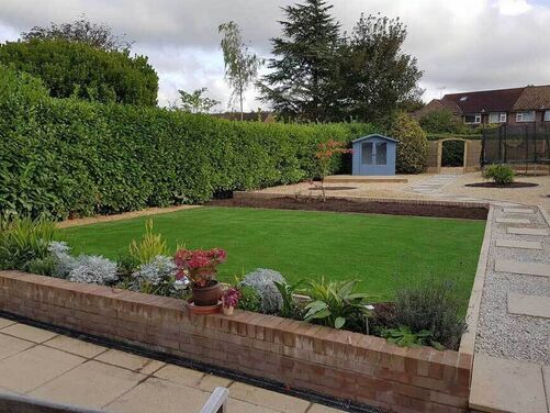 Charming backyard featuring a well-manicured lawn framed by brick flowerbeds filled with vibrant plants and shrubs. A pathway of stepping stones leads to a quaint blue garden shed, surrounded by gravel areas and lush hedging for privacy. The space is thoughtfully designed, blending functionality and aesthetics for a serene outdoor retreat