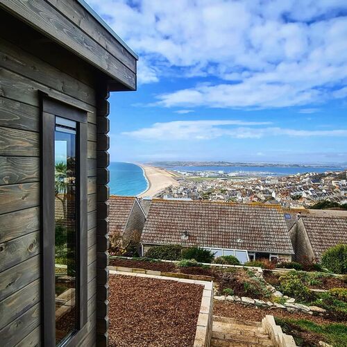 Stunning view from a timber garden building perched on a hillside, showcasing its natural wood finish and sleek modern design. The building overlooks a picturesque coastal scene, with a vibrant blue ocean, golden sandy beaches, and a charming town nestled below. Terraced landscaping with stone paths and planting beds enhances the setting, blending seamlessly with the surrounding environment. This garden building, designed and installed by Forever Timber, offers a tranquil retreat with breathtaking panoramic views, making it an ideal space for relaxation or inspiration.