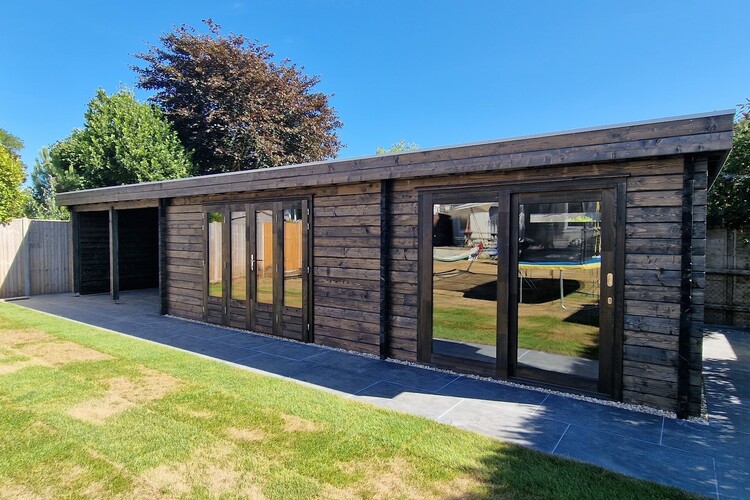 A contemporary wooden log cabin with large windows and double doors, nestled in a grassy yard. The sky is clear and blue with trees visible in the background, offering a perfect retreat for cosy accommodation.