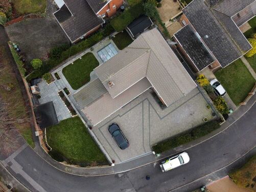 This aerial view captures a well-organized residential property with distinct landscaping and design features. The house features a pitched roof and is surrounded by a mix of paved driveways, green lawns, and carefully planned pathways. The driveway design integrates clean geometric patterns, providing a modern touch to the overall layout.

The outdoor area includes different zones for functional and aesthetic purposes, such as a small patio space, green garden areas, and accessible walkways. The surrounding neighborhood roads and adjacent homes highlight a suburban setting with ample space and attention to outdoor upkeep.

If you have specific ideas or changes you'd like to explore for this space, let me know, and I can suggest tailored landscaping or design concepts!