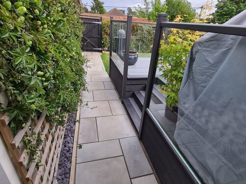 This is a beautifully designed side pathway that combines functionality with aesthetics. The pathway features clean, modern paving stones that align seamlessly with the sleek black-framed glass balustrade of the adjacent deck area. The use of a vertical trellis with climbing plants adds a natural and vibrant touch, softening the contemporary look of the hardscape.

The incorporation of slate chippings at the base of the trellis creates a neat and low-maintenance feature, ensuring that the pathway remains tidy and visually appealing. The black gate at the end provides privacy and security while blending harmoniously with the overall color scheme. This design optimizes a narrow space effectively, maintaining a balance between practicality and style.