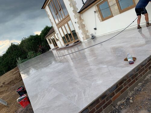 A modern house with a large, newly constructed patio featuring polished light grey tiles. The house has large glass windows and wooden-framed doors, with stone accents on the exterior. The surrounding area includes a landscaped garden with plants and an unfinished dirt section in the foreground