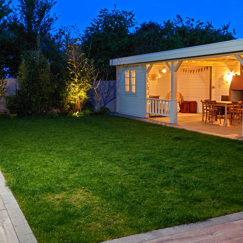 A bespoke garden gazebo supplied by Lugarde and designed and installed by Forevertimber. This spacious structure features a charming white-painted timber finish with a flat roof, supported by sturdy posts with decorative bracing. The design includes an open-sided seating and dining area, complete with warm ambient lighting, creating a cozy and inviting atmosphere for evening gatherings. A partially enclosed section with windows adds versatility and privacy. Positioned on a wooden deck adjacent to a well-maintained lawn, this gazebo seamlessly blends functionality with elegance, making it a perfect addition to a stylish outdoor living space.
