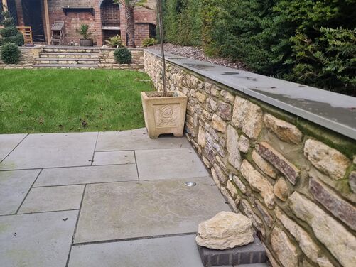 The image features a neatly landscaped garden space with a paved patio made of light gray stone slabs. A decorative stone retaining wall with natural stone and smooth coping runs along one side, adding structure and visual appeal. On the patio, a tall potted tree in a square ornamental planter adds greenery and elegance.

In the background, a raised area is visible, accessed via stone steps. This upper section includes an outdoor brick fireplace or barbecue, surrounded by shrubs, a small palm tree, and seating under a wooden pergola. Lush greenery, including dense hedges and trimmed grass, creates a serene and private atmosphere, blending formal and natural elements seamlessly.