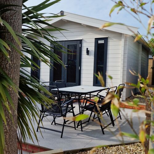 Elegant timber garden building with a pitched roof, painted in a soft white finish and complemented by contrasting dark-framed double doors and windows. Supplied by Lugarde and designed and installed by Forever Timber, the building features a modern and minimalist design ideal for a home office, studio, or relaxation space. The outdoor area includes a stylish patio with black metal dining furniture, surrounded by landscaped greenery and decorative plants, creating a serene and inviting atmosphere. The combination of high-quality craftsmanship and thoughtful design makes this structure a functional and aesthetic addition to any garden.