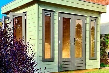Stylish modern timber garden room featuring a flat roof and painted in a soft green finish with contrasting dark gray-framed doors and windows. The structure includes large glass panels that allow ample natural light to flood the interior, creating a bright and inviting space. Designed with clean lines and a contemporary aesthetic, this garden room is ideal for use as an office, studio, or relaxation space. Surrounded by neatly landscaped greenery, it adds functionality and elegance to any outdoor area.